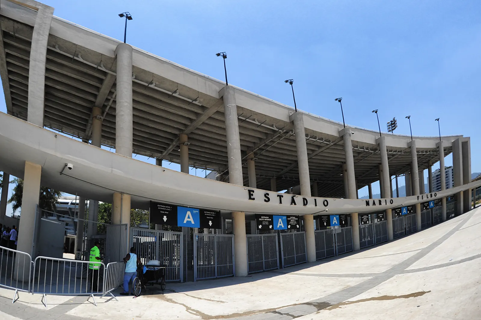 The stadium's curved concrete frontage on a sunny day, with a blue entrance sign marked A and palm trees along the plaza