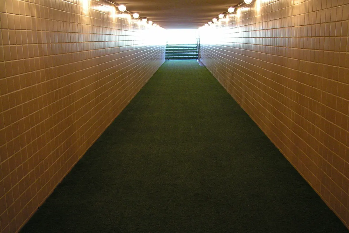 A long tunnel with orange-brown walls and a green carpet floor, lit by ceiling lamps, leading up towards daylight