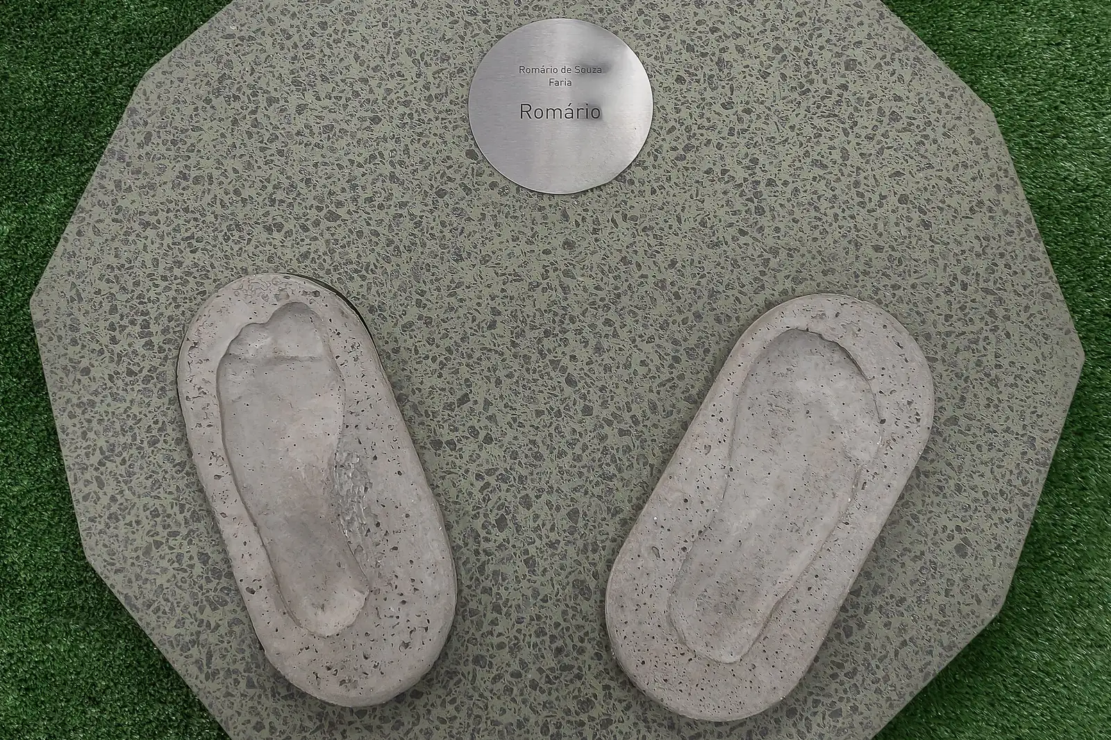 A round grey stone plaque set in grass, with a pair of footprints pressed into it and a small metal plate above them