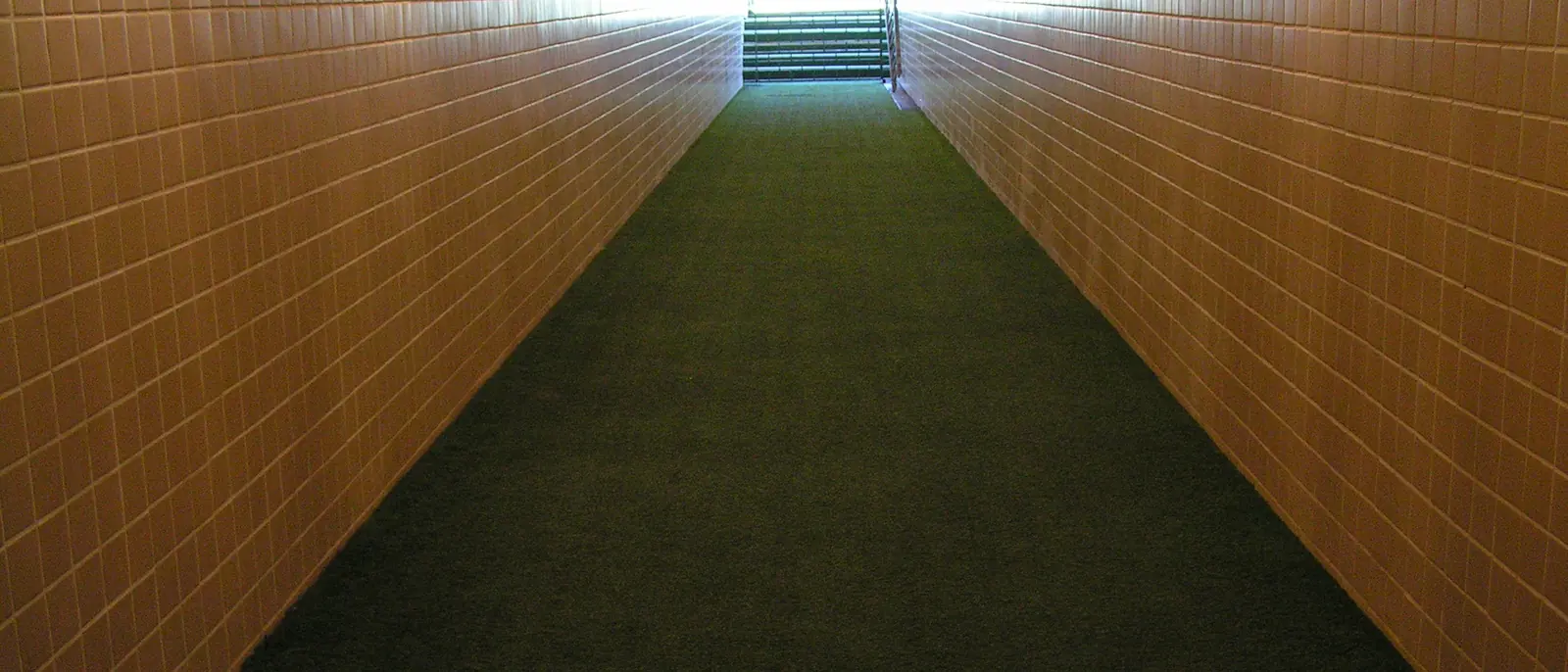 A long tunnel with orange-brown walls and a green carpet floor, lit by ceiling lamps, leading up towards daylight and a football pitch